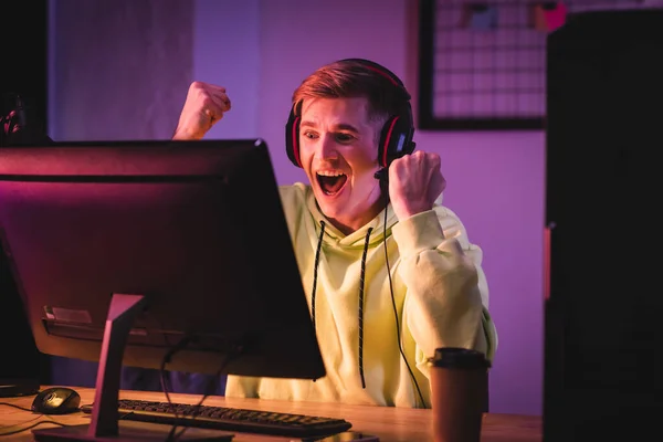 Excited gamer in headset showing yeah gesture near computer and coffee to go on blurred foreground — Stock Photo