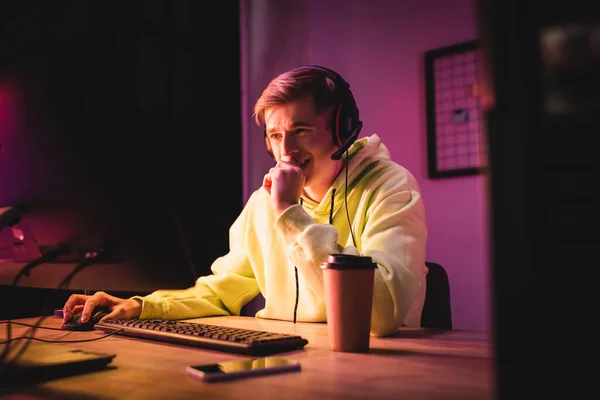 Positive gamer in headset using computer near smartphone and takeaway coffee on blurred foreground — Stock Photo