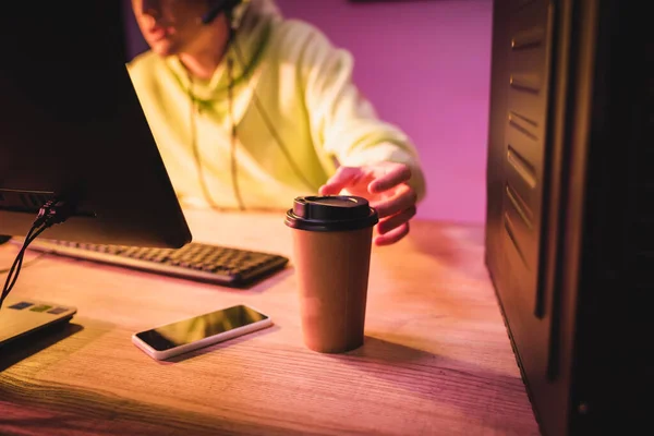 Vista ritagliata del caffè per andare e smartphone sul tavolo vicino al giocatore utilizzando il computer su sfondo sfocato — Foto stock