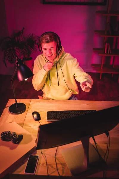 KYIV, UCRANIA - 21 de agosto de 2020: Jugador sonriente con auriculares apuntando con los dedos a la computadora cerca de joystick, caja de pizza y teléfono inteligente en primer plano borroso - foto de stock