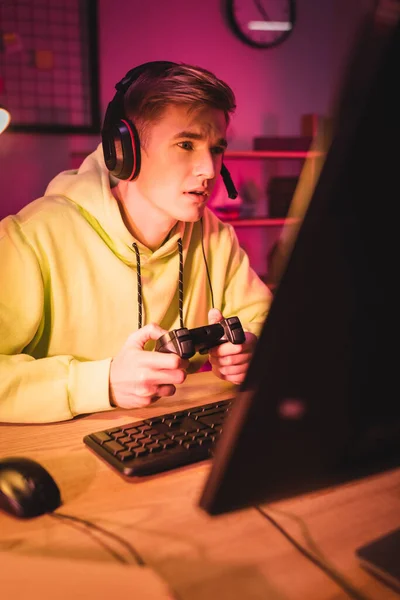 QUIIV, UCRÂNIA - 21 DE AGOSTO DE 2020: Jovem em fone de ouvido jogando videogame com joystick e computador em primeiro plano desfocado — Fotografia de Stock