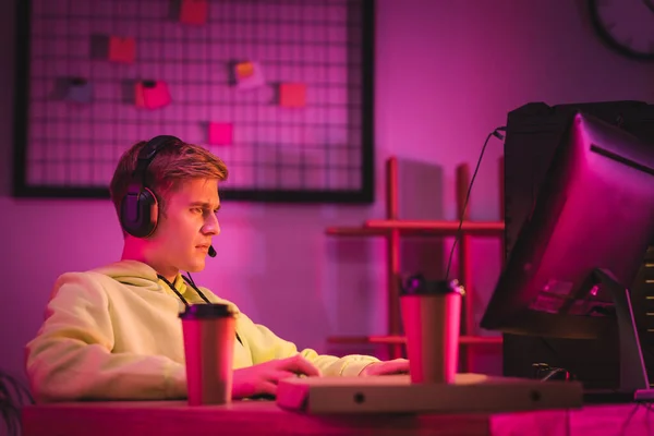 KYIV, UKRAINE - AUGUST 21, 2020: Gamer in headset looking at computer monitor near pizza box and coffee to go on blurred foreground at home — Stock Photo
