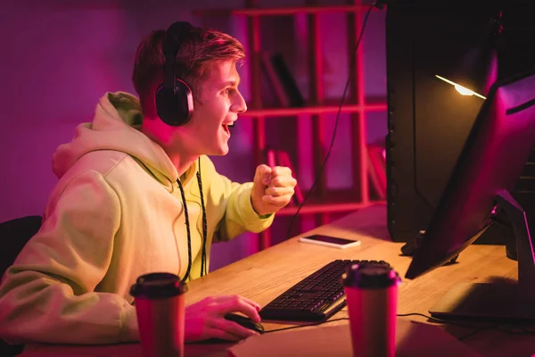 Fröhlicher Gamer zeigt Yah-Geste in der Nähe von Computer, Coffee to go und Smartphone auf verschwommenem Hintergrund — Stockfoto