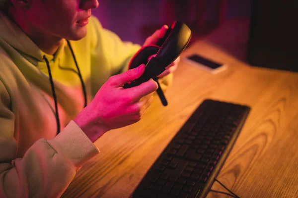 Vista recortada del gamer sosteniendo auriculares cerca del teclado de la computadora sobre fondo borroso - foto de stock