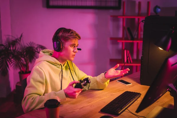 KYIV, UKRAINE - AUGUST 21, 2020: Gamer in headset holding joystick while pointing at computer monitor near smartphone and coffee to go on blurred foreground — Stock Photo