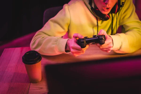 KYIV, UCRANIA - 21 de agosto de 2020: Vista recortada del joystick en las manos del jugador emocionado cerca del café para ir y monitor de computadora en primer plano borroso - foto de stock