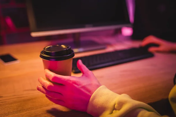 Обрезанный вид человека, принимающего кофе, чтобы пойти, играя в видеоигру на компьютере на размытом фоне — стоковое фото