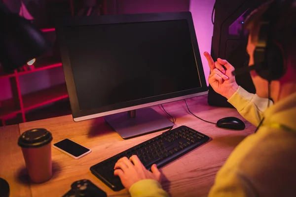 Jogador no fone de ouvido mostrando o dedo médio no monitor do computador enquanto joga jogo de vídeo perto do smartphone e café takeaway em primeiro plano desfocado — Fotografia de Stock