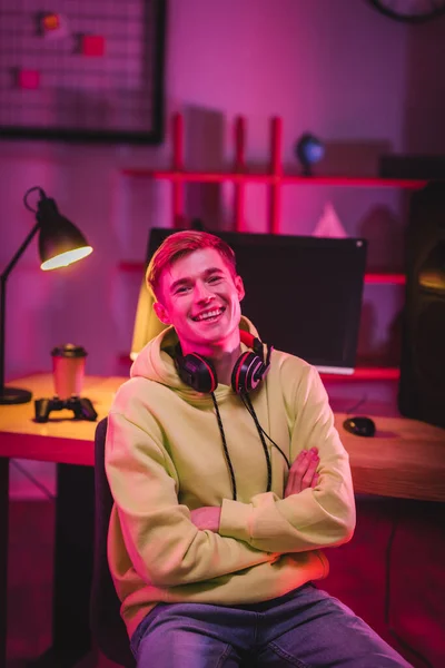 Jugador sonriente en auriculares mirando a la cámara mientras está sentado cerca de la computadora y el mando sobre un fondo borroso — Stock Photo