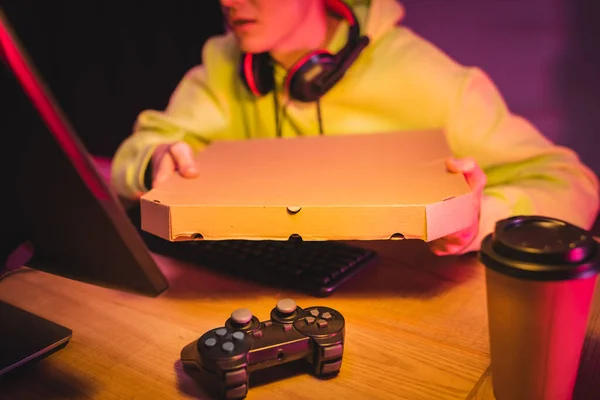KYIV, UKRAINE - AUGUST 21, 2020: Joystick and coffee to go near gamer in headphones holding pizza box near computer on blurred background — Stock Photo