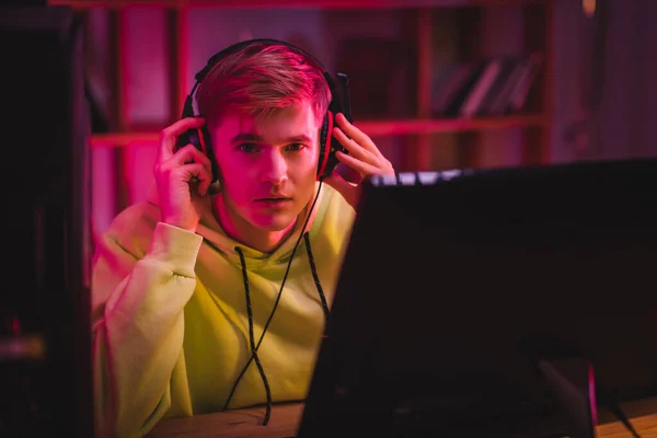 Jovem jogador segurando fones de ouvido e olhando para a câmera perto do monitor do computador em primeiro plano desfocado — Fotografia de Stock