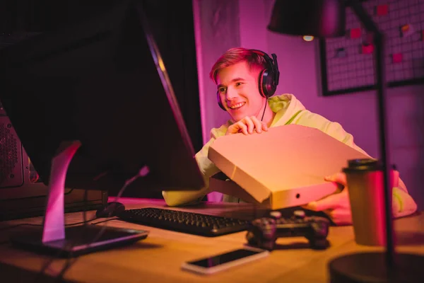 Jugador sonriente en auriculares que sostiene la caja de pizza cerca de la computadora, el joystick y el teléfono inteligente en primer plano borroso - foto de stock