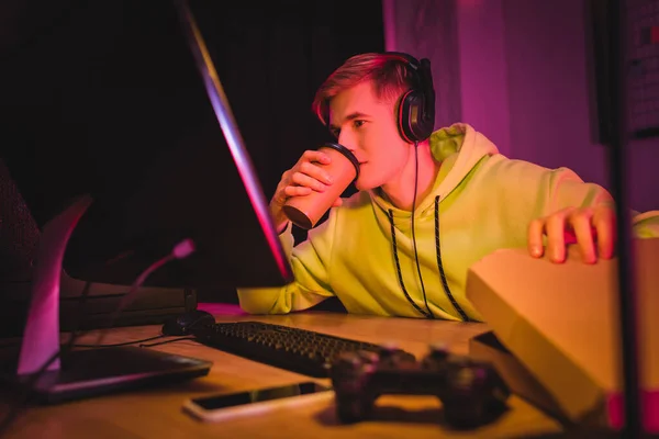 Joven jugador en auriculares bebiendo café para ir y sosteniendo la caja de pizza cerca de la computadora y el joystick en primer plano borroso — Stock Photo