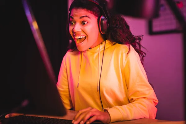 Alegre jogador americano africano no fone de ouvido jogando videogame no computador em primeiro plano desfocado — Fotografia de Stock