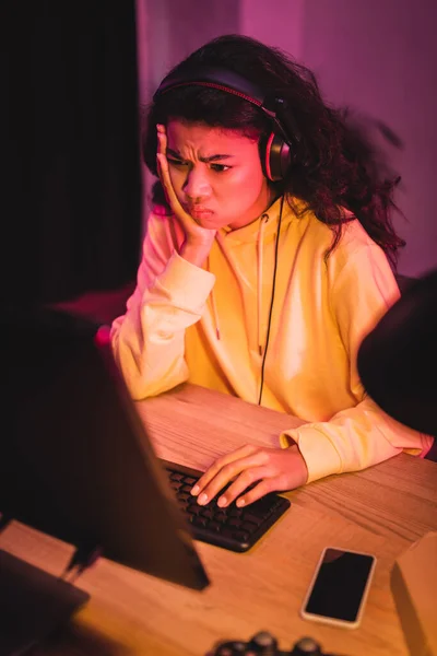 Perturbado jugador afroamericano en auriculares mirando el monitor de la computadora cerca del teléfono inteligente en primer plano borroso - foto de stock