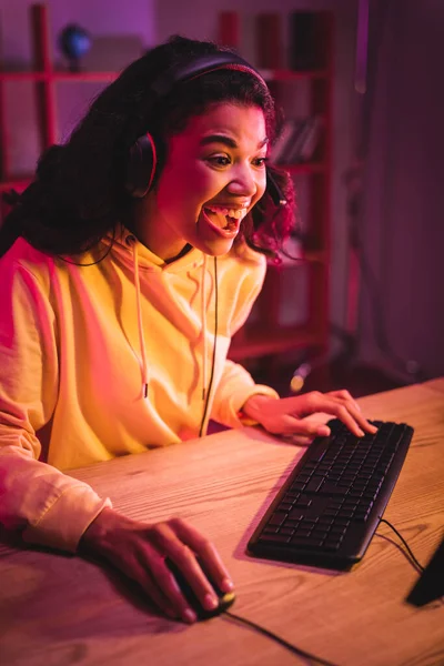 Excited african american gamer in headset using computer keyboard and mouse on blurred foreground — Stock Photo