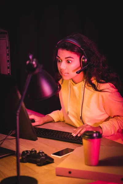 KIEW, UKRAINE - 21. AUGUST 2020: Aufgeregter afrikanisch-amerikanischer Gamer im Headset mit Computer in der Nähe von Steuerknüppel, Smartphone und Pizzakiste auf verschwommenem Vordergrund — Stockfoto