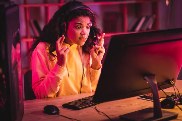 KYIV, UKRAINE - AUGUST 21, 2020: Excited african american gamer in headset sitting with crossed fingers near computer, joystick and smartphone at home — Stock Photo