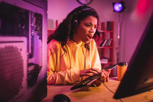 Jugador afroamericano insatisfecho en auriculares usando el teclado de la computadora cerca del café para ir - foto de stock