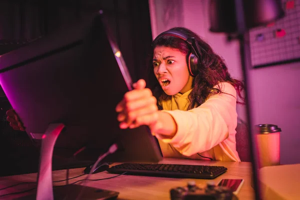 Crazy african american gamer in headset holding computer monitor near coffee to go, smartphone and joystick on blurred foreground — Stock Photo
