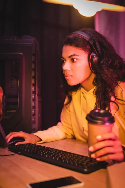 African american player in headset playing video game on computer and holding takeaway coffee on blurred foreground — Stock Photo