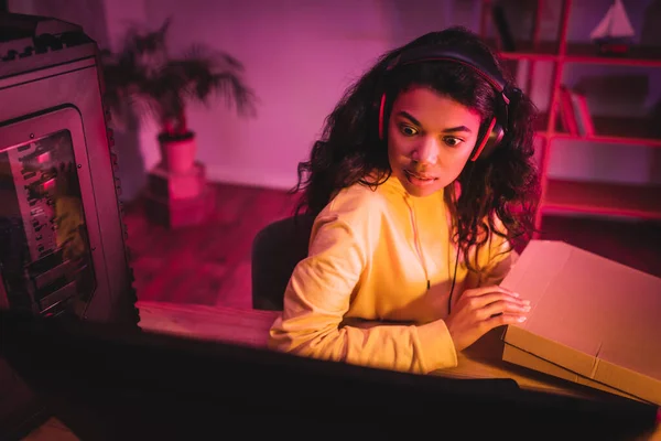 African american gamer in headset holding pizza box and looking at computer monitor on blurred foreground — Stock Photo