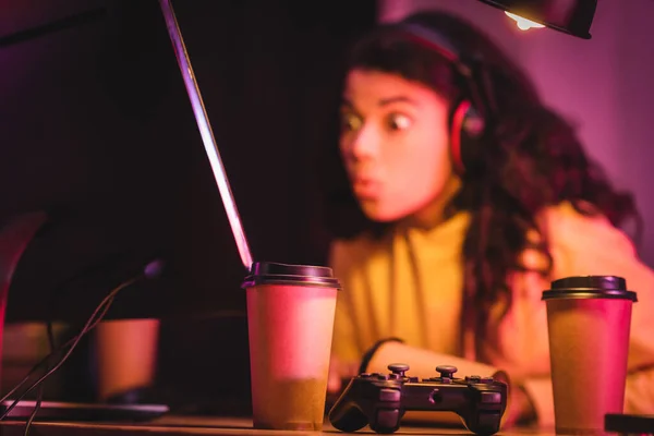 KYIV, UKRAINE - AUGUST 21, 2020: Joystick and coffee to go near excited african american gamer and computer on blurred background — Stock Photo
