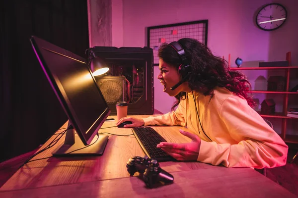 KYIV, UKRAINE - 21 DE AGOSTO DE 2020: Jogador afro-americano irritado no fone de ouvido usando o computador perto do gamepad e café para ir — Fotografia de Stock