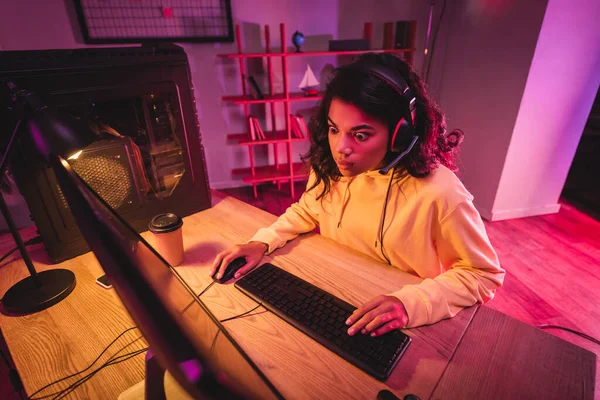 Sorprendido jugador afroamericano en auriculares usando la computadora durante el videojuego cerca del café para ir - foto de stock