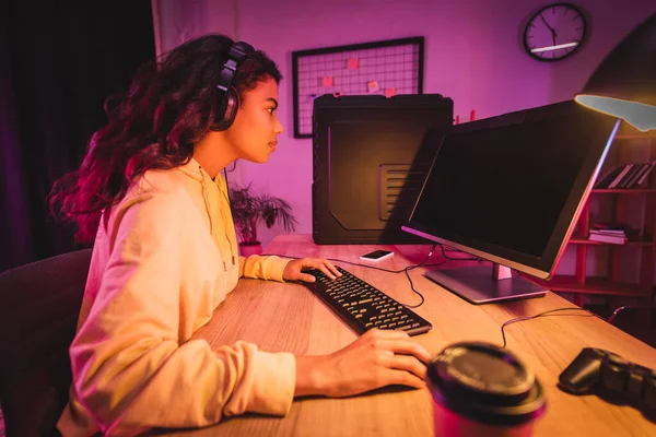 Vista lateral do jogador americano africano no fone de ouvido jogando videogame no computador com tela em branco perto de joystick e café para ir em primeiro plano desfocado — Stock Photo