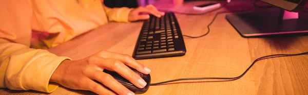 Vista recortada de la mujer usando el ratón de la computadora mientras que juega el videojuego en fondo borroso, bandera - foto de stock