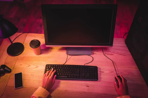 Vista recortada del jugador utilizando el ordenador cerca de teléfono inteligente, café para llevar y gamepad en la mesa - foto de stock