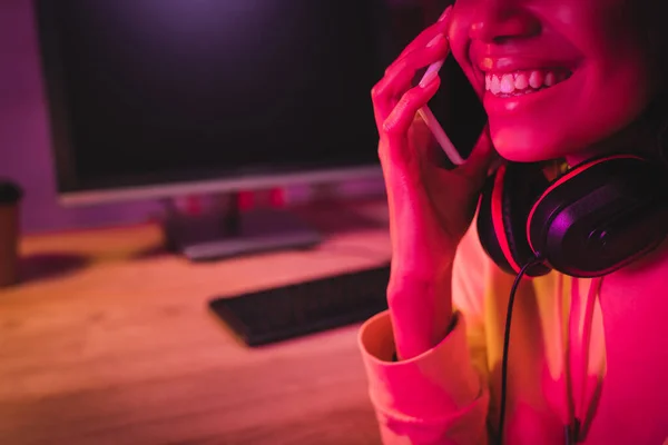 Vista cortada de mulher americana africana sorridente em fones de ouvido falando no smartphone perto do computador no fundo embaçado — Fotografia de Stock