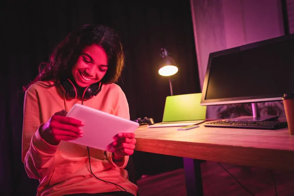 Усміхнений афроамериканський геймер в гарнітурі, використовуючи цифровий планшет біля комп'ютерів і кави, щоб йти на розмитому фоні — стокове фото