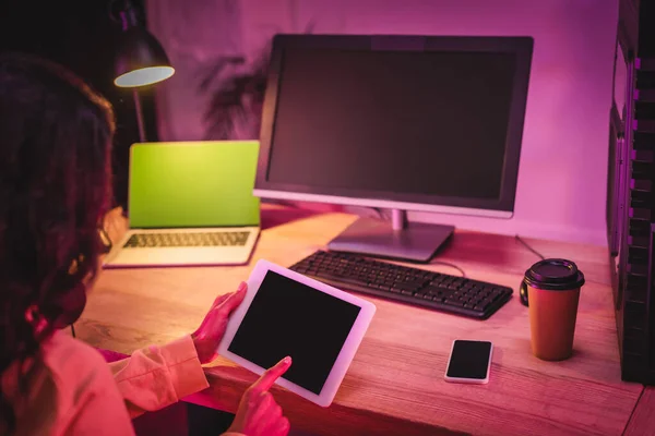 Digital tablet with blank screen in hands of gamer near computers, smartphone and coffee to go on blurred background — Stock Photo