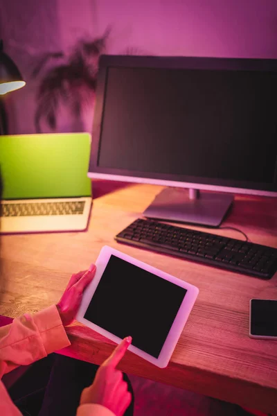 Vista recortada de una mujer sosteniendo una tableta digital con pantalla en blanco cerca de un teléfono inteligente y computadoras en un fondo borroso - foto de stock