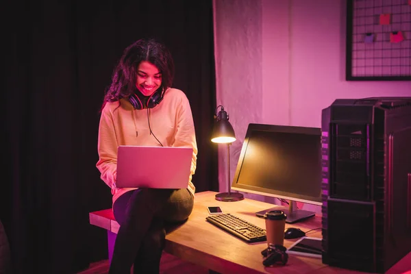 Cheerful african american gamer in headset using laptop near devices, gamepad and coffee to go on blurred foreground — Stock Photo