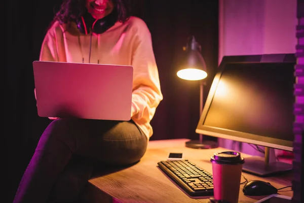 Vista ritagliata di sorridente giocatore afroamericano utilizzando laptop vicino al caffè per andare e dispositivi in primo piano sfocato — Stock Photo