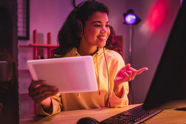 Sonriente jugador afroamericano en auriculares apuntando con el dedo a la computadora mientras sostiene la tableta digital en primer plano borroso - foto de stock