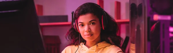 Africano jugador americano en auriculares mirando la cámara cerca de la computadora en primer plano borrosa, bandera - foto de stock