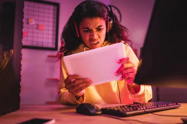 Arrabbiato giocatore afroamericano in auricolare utilizzando tablet digitale vicino al computer in primo piano offuscata — Foto stock