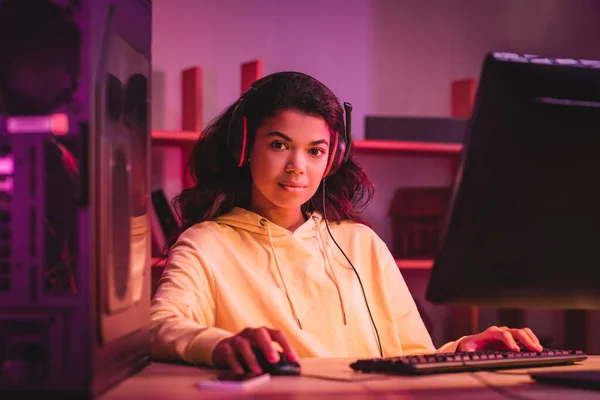 Africano jugador americano en auriculares mirando a la cámara cerca de la computadora en primer plano borrosa - foto de stock