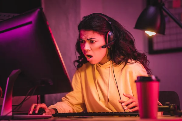 Jogador afro-americano chocado em fone de ouvido usando o computador perto de café para ir e joystick em primeiro plano desfocado — Fotografia de Stock