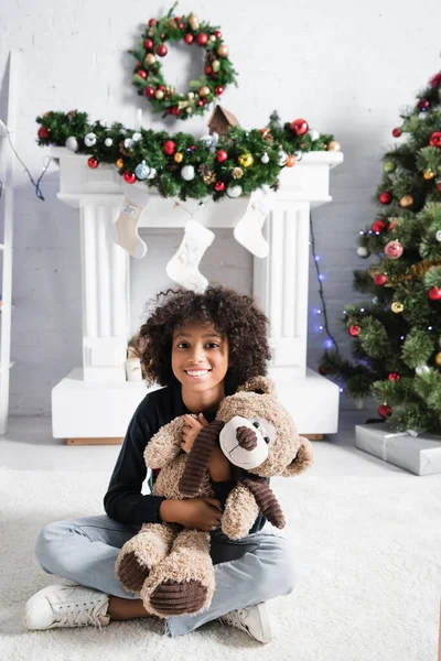 Lächelnde afrikanische Amerikanerin blickt in die Kamera, während sie mit Teddybär auf verschwommenem Hintergrund mit Weihnachtsdekoration auf dem Boden sitzt — Stockfoto