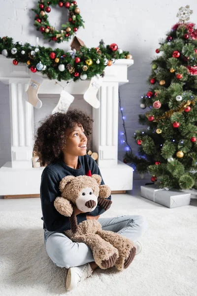 Glücklich afrikanisch-amerikanisches Mädchen schaut weg, während es mit Teddybär auf dem Boden in der Nähe des Weihnachtsbaums sitzt — Stockfoto