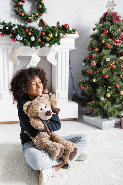 Aufgeregtes afrikanisches amerikanisches Mädchen sitzt mit geschlossenen Augen auf dem Boden und umarmt Teddybär in der Nähe von Kamin und Weihnachtsbaum — Stockfoto
