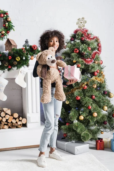 Happy african american girl holding gift box and teddy bear near decorated christmas tree and fireplace — Stock Photo