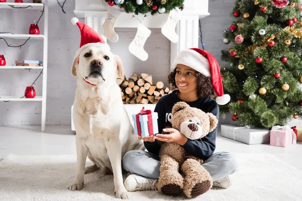 Feliz afroamericana chica sosteniendo caja de regalo y osito de peluche mientras está sentado en el suelo cerca de perro labrador sobre fondo borroso - foto de stock