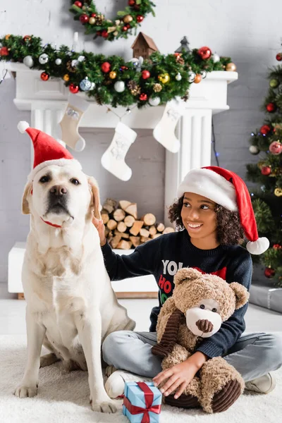 Cheerful african american girl stroking labrador while sitting on floor with teddy bear near gift box and decorated fireplace on blurred background — Stock Photo