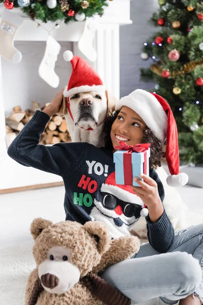 Smiling african american girl stroking labrador while sitting on floor with gift box and teddy bear on blurred background — Stock Photo
